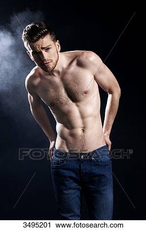 Stock Image - Topless man, black background smoke behind; wearing jeans, serious, forehead creased, hands in jeans backpockets. Fotosearch