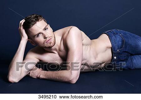 Stock Image - Topless man, dark blue background; lying on the floor, holding his head with his hand, wearing jeans, languid, looking at the camera. Fotosearch