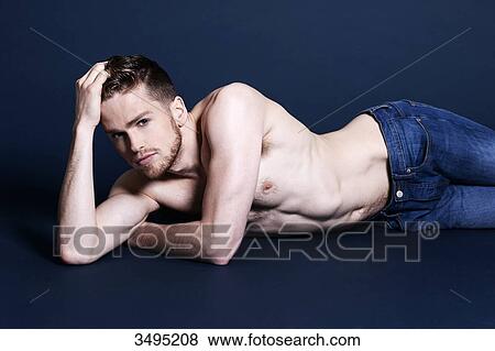 Stock Photo - Topless man, dark blue background; lying on the floor, serious, one hand in his hair, holding his head with his hand, wearing jeans, looking at the camera. Fotosearch