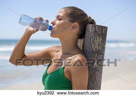 Stock Image - Woman leaning against a wooden post on the beach drinking water. Fotosearch