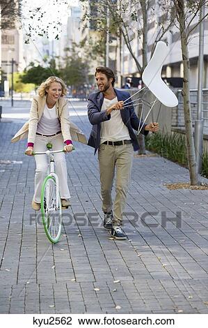 Stock Image - Woman riding a bicycle with a man carrying a chair beside her. Fotosearch