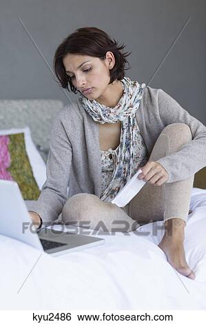 Stock Photograph - Woman working on a laptop with holding a paper on the bed. Fotosearch