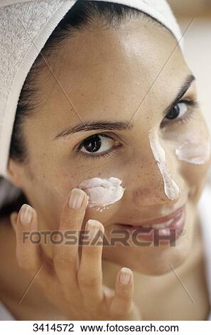 Stock Image - Close-up of a smiling, dark haired young woman applying face cream. Fotosearch