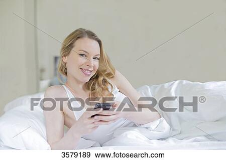 Stock Photo - Content pretty blonde woman with mobile in bed smiling at camera. Fotosearch