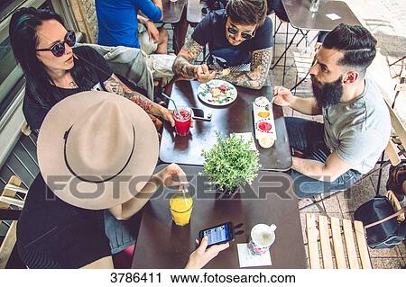 Stock Image - Group of young hipster sitting in a restaurant terrace, eating and drinking. Fotosearch