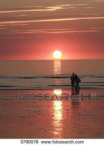Stock Photo - Normandy, couple with dog in the background looking at the sunset on a beach. Fotosearch