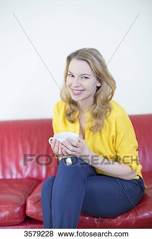 Stock Photo - Portrait of a beautiful blonde woman enjoying a cup of tea on the sofa and smiling at camera. Fotosearch