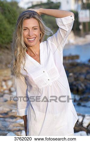 Stock Image - Portrait of a smiling young blonde woman in white shirt on the rock in the sea.. Fotosearch