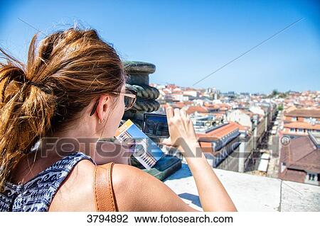 Stock Image - Portrait of a young woman from behind, taking a picture with his smartphone on the heights of a batiement the Commerce Square, Lisbon, Portugal. Fotosearch