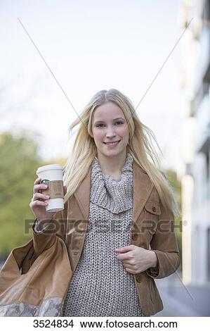 Picture - Pretty blonde woman with cafe to go walking in city center and smiling at camera. Fotosearch