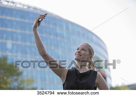 Pretty young woman taking a selfie in front of an Office Building View Large Photo Image Picture - Pretty young woman taking a selfie in front of an Office Building. Fotosearch