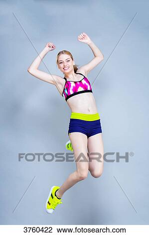Stock Image - Smiling young woman dressed in fitness, front, jumps on a gray background. Fotosearch