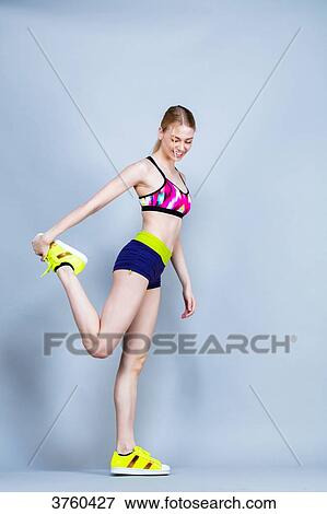Stock Photo - Smiling young woman dressed in fitness, profile, holding his foot on a gray background. Fotosearch