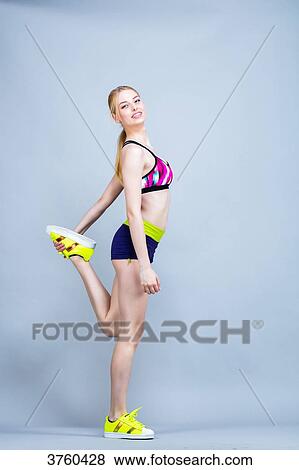 Stock Photo - Smiling young woman dressed in fitness, profile, holding his foot on a gray background. Fotosearch