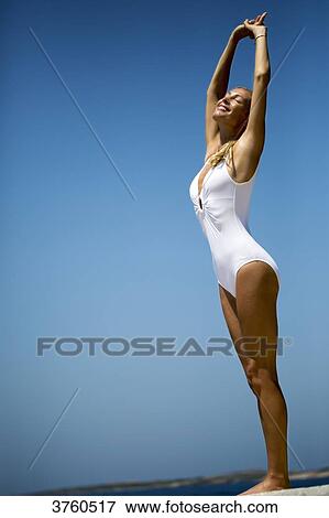 Tanned young woman in white swimsuit streching arms in the air facing a blue sky. View Large Photo Image Stock Photo - Tanned young woman in white swimsuit streching arms in the air facing a blue sky.. Fotosearch