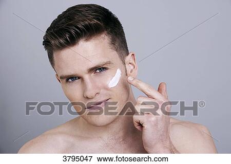 Stock Photo - topless man, looking at camera, serious, applying cream on his cheek with his finger. Fotosearch