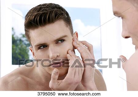 Stock Image - topless man, looking in the mirror, looking at his face in the mirror, looking at his eye wrinkles, window in the background, back of the shoulder blur on the foreground. Fotosearch