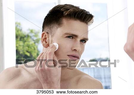 Stock Image - topless man, looking in the mirror, cleaning his face with a cotton pad, window in the background, back of the shoulder blur on the foreground, serious. Fotosearch