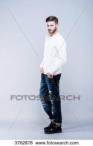 Young man standing in white sweater and blue jeans, face, hands in pockets, looking toward the lens View Large Photo Image Stock Photograph - Young man standing in white sweater and blue jeans, face, hands in pockets, looking toward the lens. Fotosearch