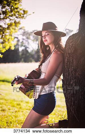 Young woman back to a tree plays guitar View Large Photo Image Picture - Young woman back to a tree plays guitar. Fotosearch