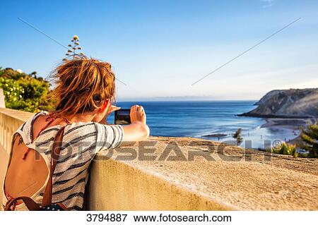 Young woman from behind, takes a photo with his smartphone on a low wall from the beach of Burgau, Algarve region, Portugal View Large Photo Image Stock Photo - Young woman from behind, takes a photo with his smartphone on a low wall from the beach of Burgau, Algarve region, Portugal. Fotosearch