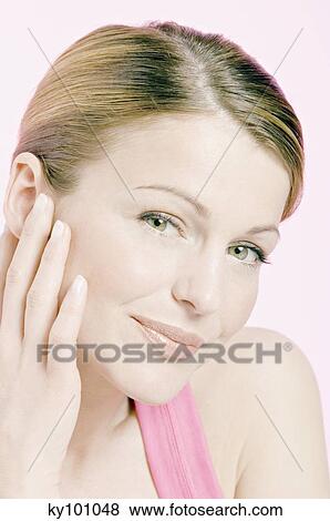 Stock Photo - Young Woman face with make up, looking at the camera, smiling, hand on her cheek, close-up (studio). Fotosearch