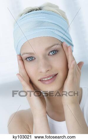 Stock Image - Young Woman face looking at the camera, putting her hands on her cheeks, close-up (studio). Fotosearch
