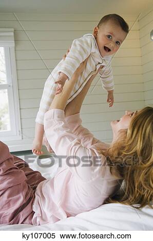 Young woman lying on the bed, holding her baby, laughing View Large Photo Image Stock Photography - Young woman lying on the bed, holding her baby, laughing. Fotosearch