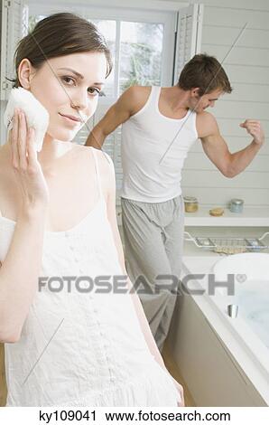 Couple in a bathroom, woman cleansing face with cotton wool, man flexing muscles in background View Large Photo Image Stock Image - Couple in a bathroom, woman cleansing face with cotton wool, man flexing muscles in background. Fotosearch