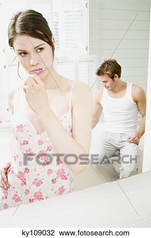 Stock Image - Young woman in nightgown brushing her teeth in front of bathroom mirror, man sitting on tub edge in background. Fotosearch