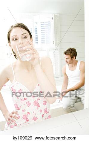 Young woman in nightgown drinking water in front of bathroom mirror, man sitting on tub edge in background View Large Photo Image Stock Photo - Young woman in nightgown drinking water in front of bathroom mirror, man sitting on tub edge in background. Fotosearch