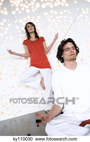 Couple in yoga position, man sitting cross-legged, woman standing in background View Large Photo Image Stock Image - Couple in yoga position, man sitting cross-legged, woman standing in background. Fotosearch