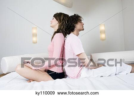 Stock Photo - Couple sitting on a bed, back to back, wearing earphones. Fotosearch