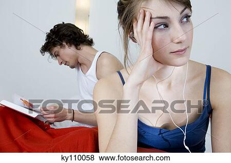 Couple sitting on a bed, thinking woman wearing headphones, man reading in background View Large Photo Image Stock Photo - Couple sitting on a bed, thinking woman wearing headphones, man reading in background. Fotosearch