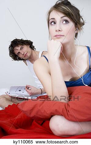 Couple sitting on a bed, thinking woman wearing headphones, man reading in background View Large Photo Image Stock Photography - Couple sitting on a bed, thinking woman wearing headphones, man reading in background. Fotosearch