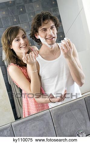 Stock Photography - Man applying shaving cream on his face in front of bathroom mirror, smiling woman resting on his shoulder. Fotosearch