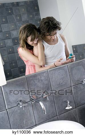 Stock Photograph - Man with shaving cream in his hand in front of bathroom mirror, smiling woman resting on his shoulder. Fotosearch