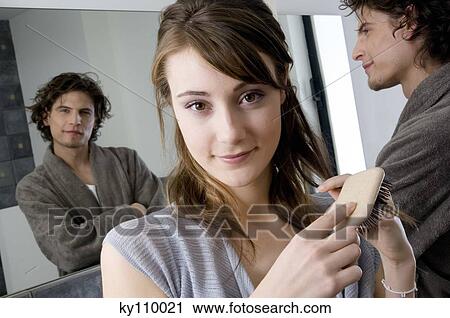 Portrait young woman brushing her hair, looking at the camera, man in bathrobe in front of bathroom mirror in background View Large Photo Image Stock Image - Portrait young woman brushing her hair, looking at the camera, man in bathrobe in front of bathroom mirror in background. Fotosearch