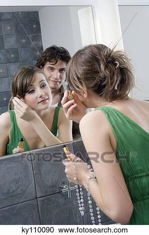 Woman applying mascara in front of bathroom mirror, man in background View Large Photo Image Stock Image - Woman applying mascara in front of bathroom mirror, man in background. Fotosearch