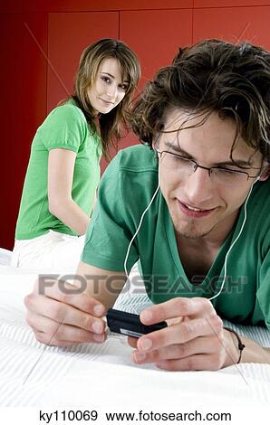 Young man lying on bed, listening to MP3 player, woman sitting in background View Large Photo Image Stock Photo - Young man lying on bed, listening to MP3 player, woman sitting in background. Fotosearch