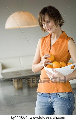 Picture - Young woman holding a bowl of oranges, looking at the camera. Fotosearch