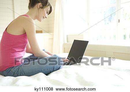 Young woman sitting leg-crossed on a bed, using laptop computer View Large Photo Image Stock Photo - Young woman sitting leg-crossed on a bed, using laptop computer. Fotosearch