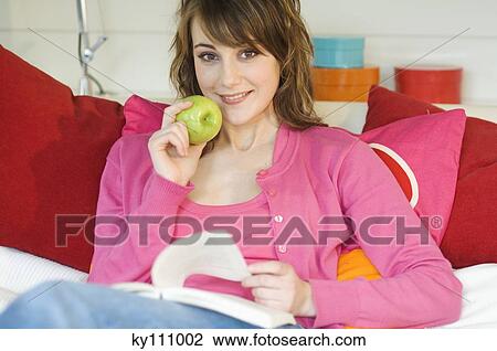 Young woman sitting on a sofa reading, holding an apple View Large Photo Image Stock Image - Young woman sitting on a sofa reading, holding an apple. Fotosearch
