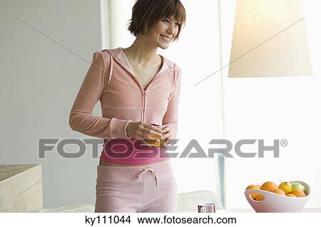 Young woman standing in kitchen, holding glass of orange juice View Large Photo Image Picture - Young woman standing in kitchen, holding glass of orange juice. Fotosearch