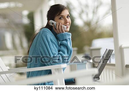 Picture - Young woman using mobile phone, sitting on a chair, looking at the camera, outdoors. Fotosearch