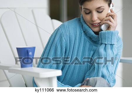 Stock Photograph - Young woman using mobile phone, sitting on a chair, outdoors. Fotosearch