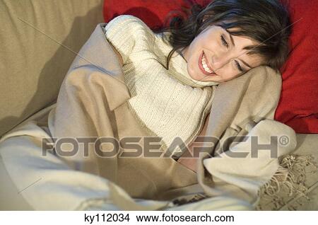 Picture - Young woman lying on a sofa, smiling, wrapped in blanket. Fotosearch