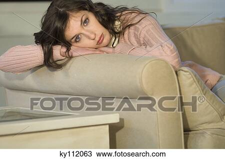 Stock Image - Young woman resting head on sofa armrest, looking at the camera. Fotosearch