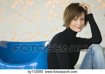 Stock Image - Young woman sitting on the floor, leaning against blue armchair, looking at the camera. Fotosearch