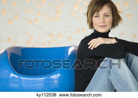 Stock Photo - Young woman sitting on the floor, leaning against blue armchair. Fotosearch
